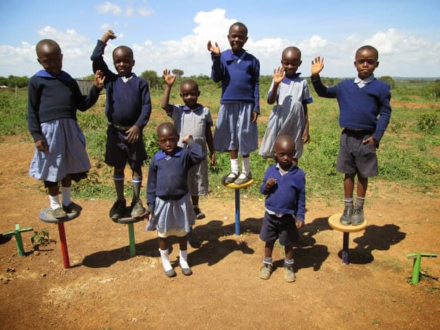 Children in school uniforms standing on small playground stools, waving
